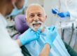 Smiling elderly man undergoing a dental checkup while a dentist uses tools to examine his teeth in a modern clinic.
